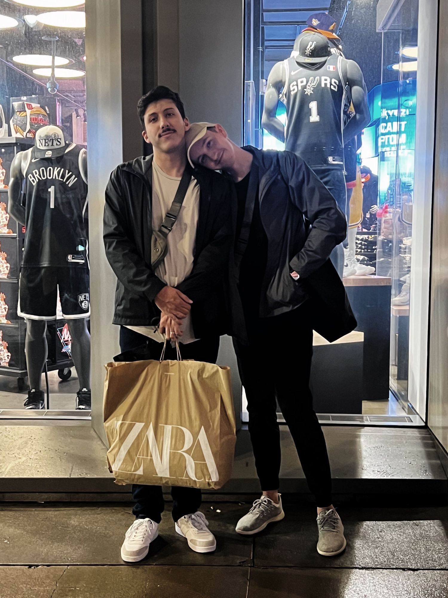 Two friends pose outside an NBA store holding a Zara shopping bag, with Brooklyn Nets and Spurs jerseys displayed behind them.