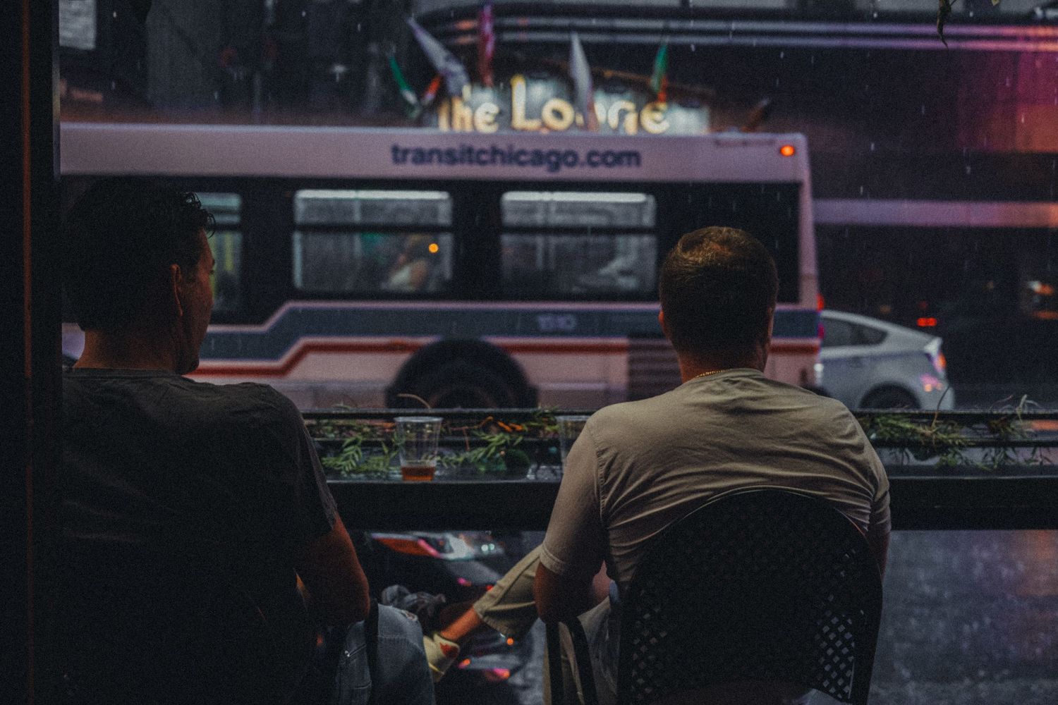 Two men sit with their backs to camera at night, watching a Chicago Transit Authority bus pass by.