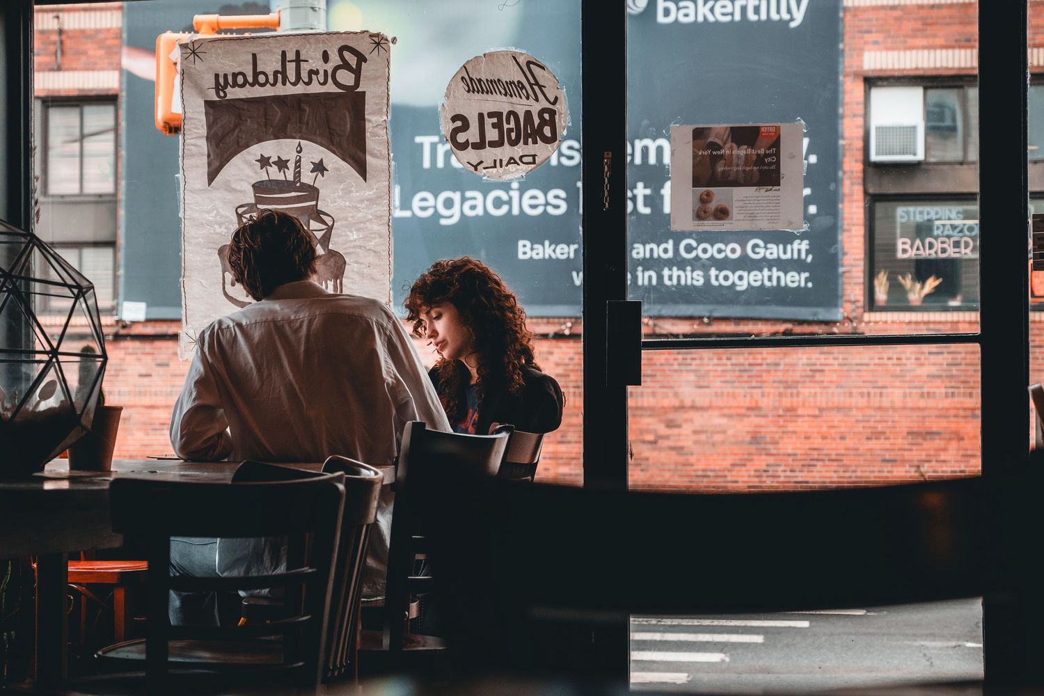 Two people sit together at a cafe table near large windows with urban street reflections and bakery signage visible.