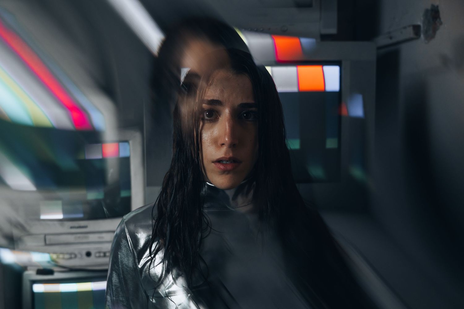 A woman with dark hair in a chrome bodysuit poses in a dimly lit room with vintage monitors and colorful light streaks.