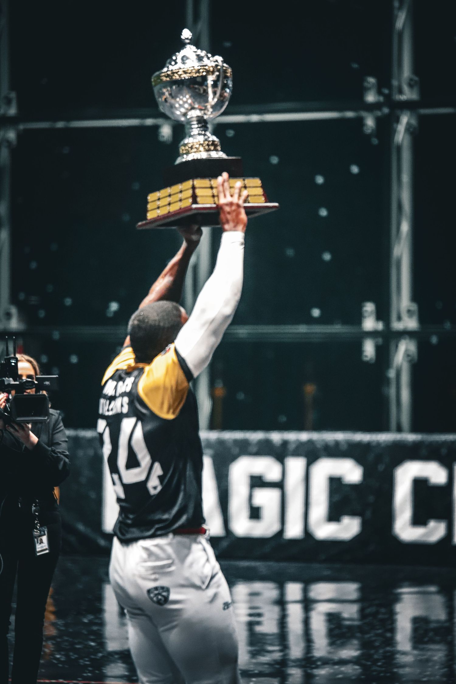 Jai Alai player wearing black and yellow uniform lifting a championship trophy celebrating on the court.