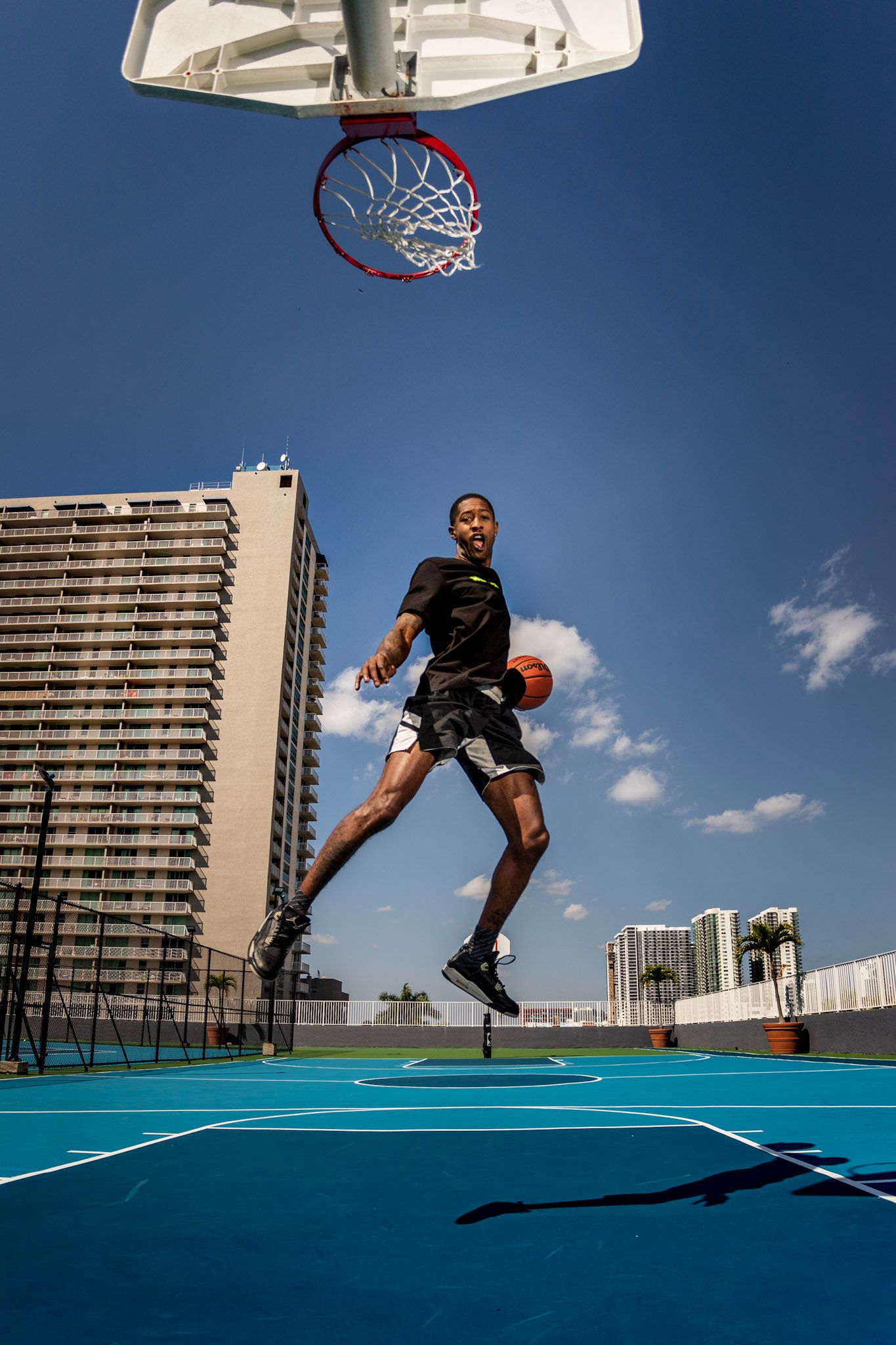 Young male basketball player in black shirt dunking while hanging on the rim on a rooftop basketball court on a sunny day
