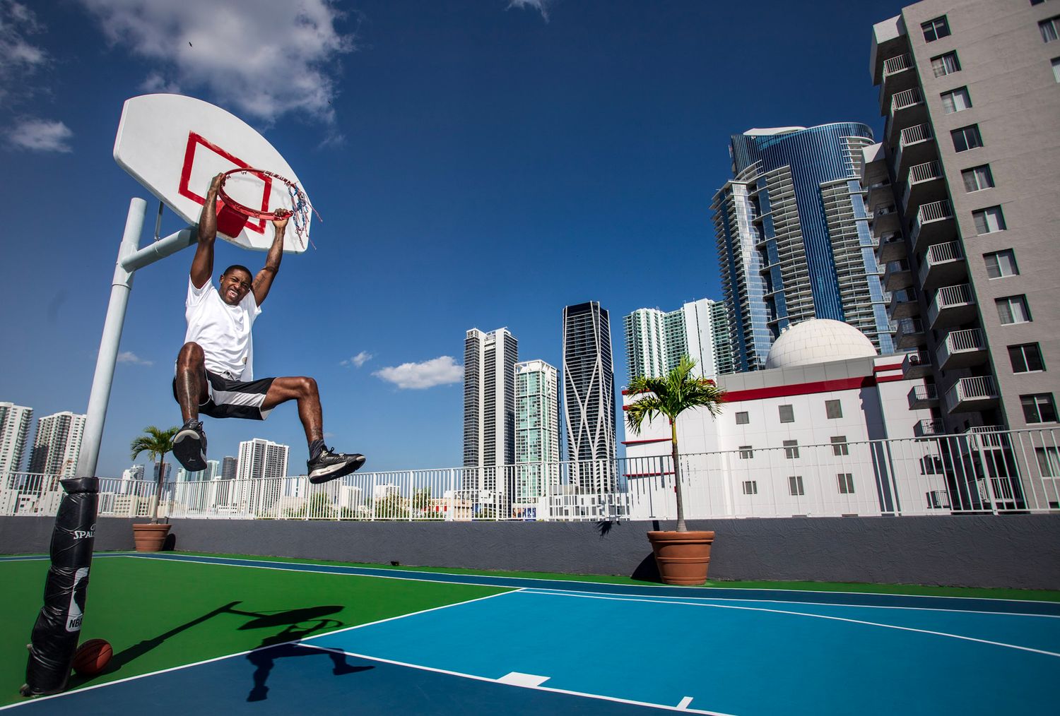 Young male basketball player in white shirt dunking while hanging on the rim on a rooftop basketball court on a sunny day