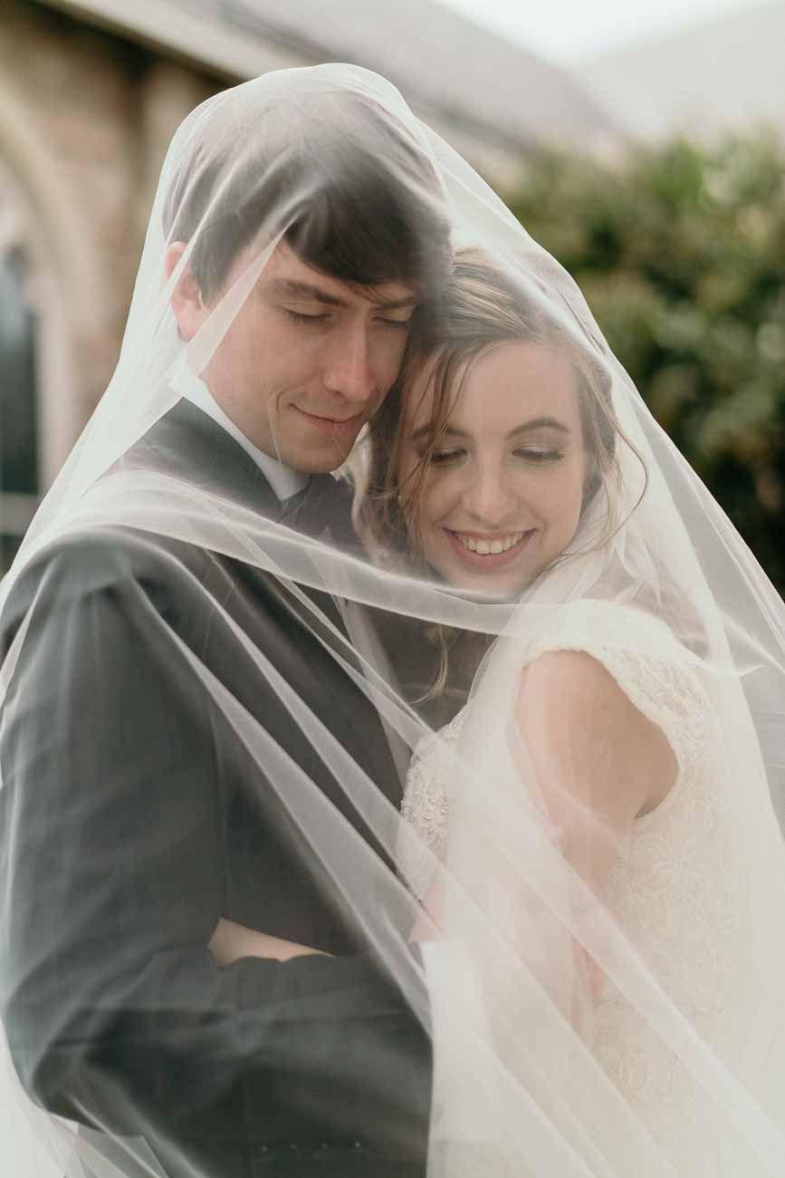 Bride smiles as groom embraces her tenderly beneath her flowing veil outside a stone building.