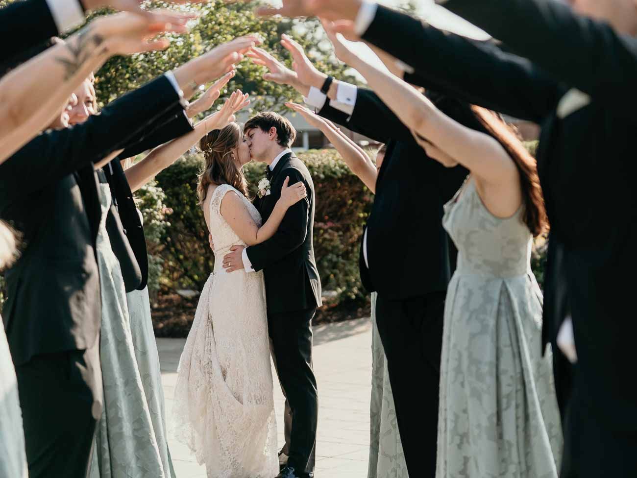 Wedding party forms an arch with their arms as bride and groom share a romantic kiss beneath.