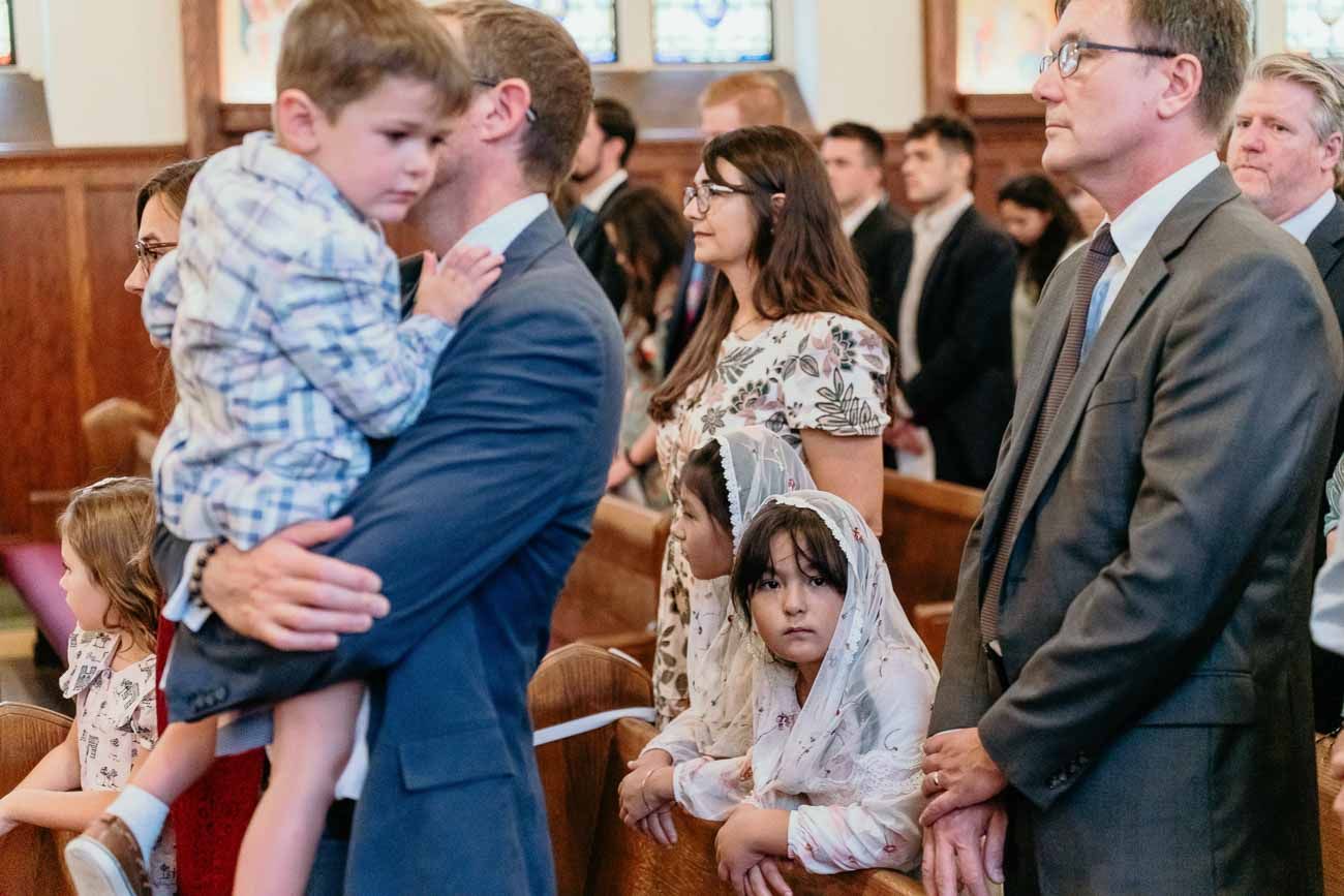 Father holds young child while family members stand together in church pews during wedding.