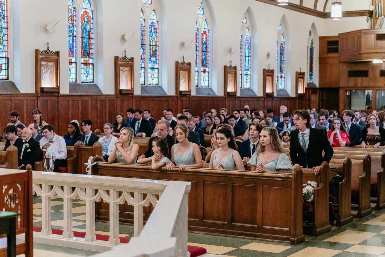 Wedding guests fill wooden pews of a beautiful Catholic church with tall stained glass windows.