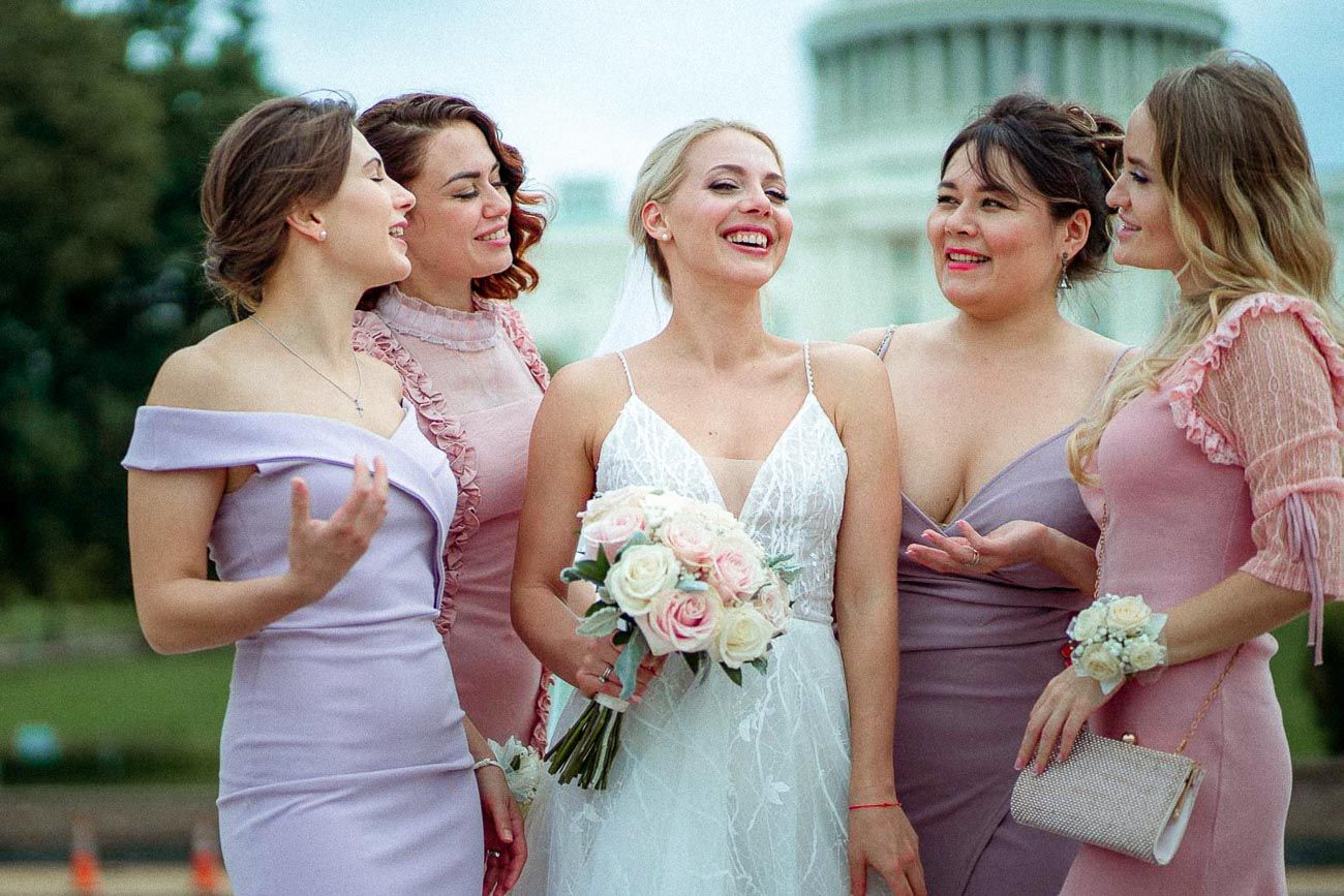 Bride in white lace gown laughing with four bridesmaids in pink dresses near the US Capitol.