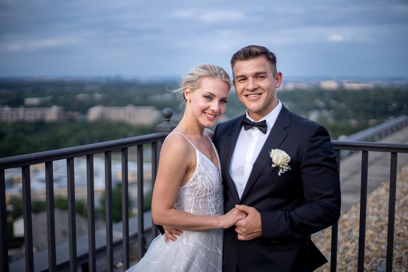Smiling newlywed couple posing on a rooftop terrace with a panoramic city skyline view in the background.