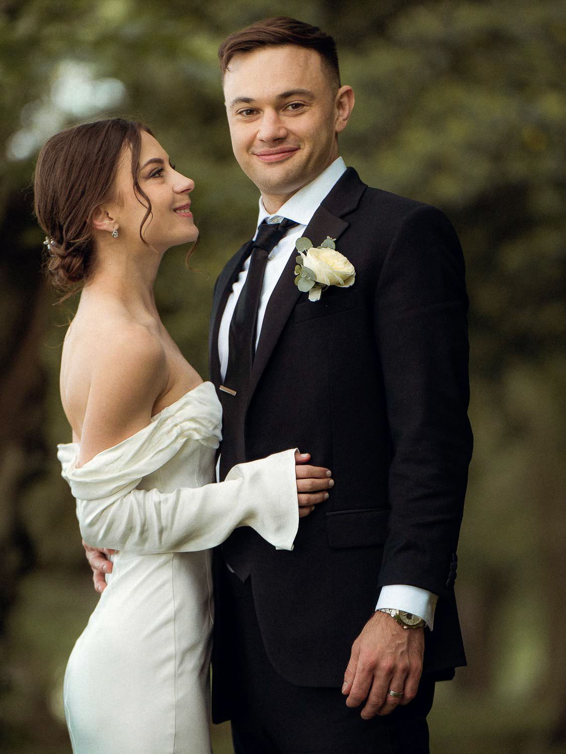 Bride in off-shoulder white gown and groom in black suit pose together in a green park.
