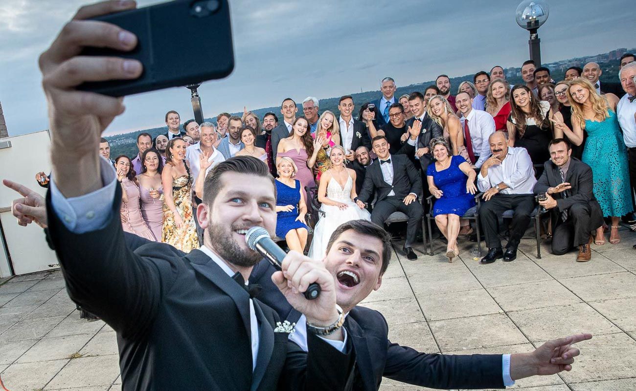 Two men take selfie with large wedding party crowd gathered behind them outdoors.