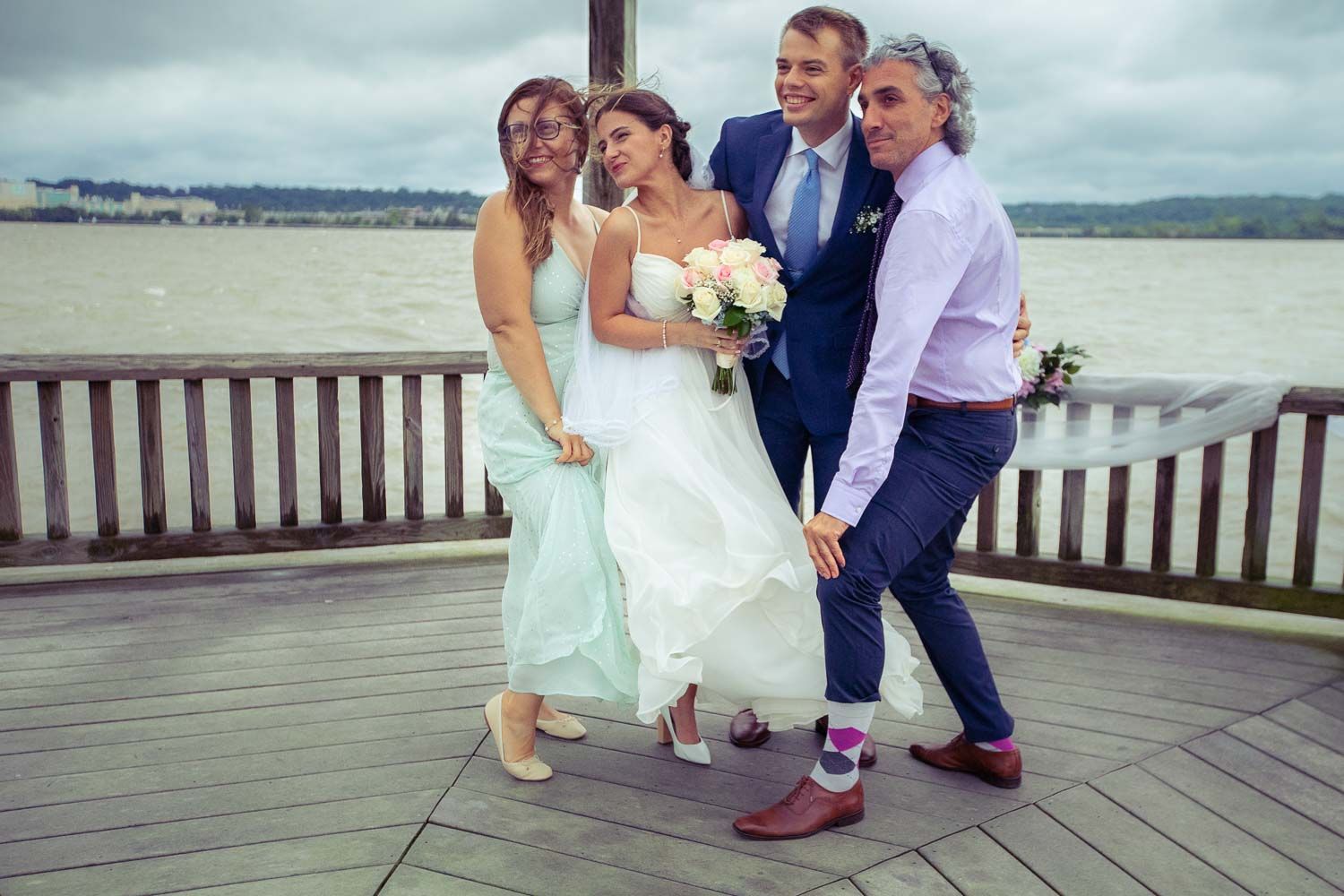 Happy wedding party of four posing together on a waterfront deck on a cloudy day.