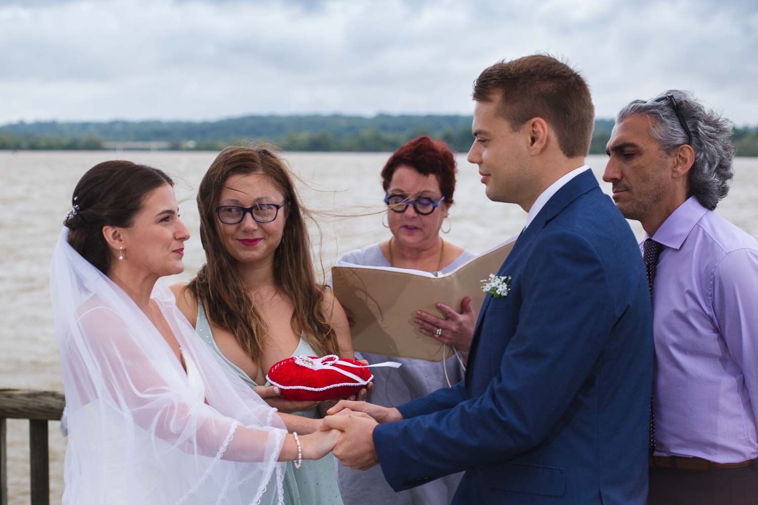 Groom places wedding ring on brides finger during emotional outdoor waterfront ceremony with guests watching.