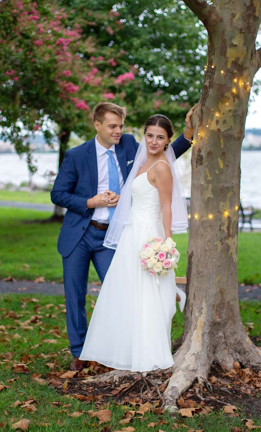 Bride and groom posing by a tree with fairy lights in a park near a waterfront on their wedding day.
