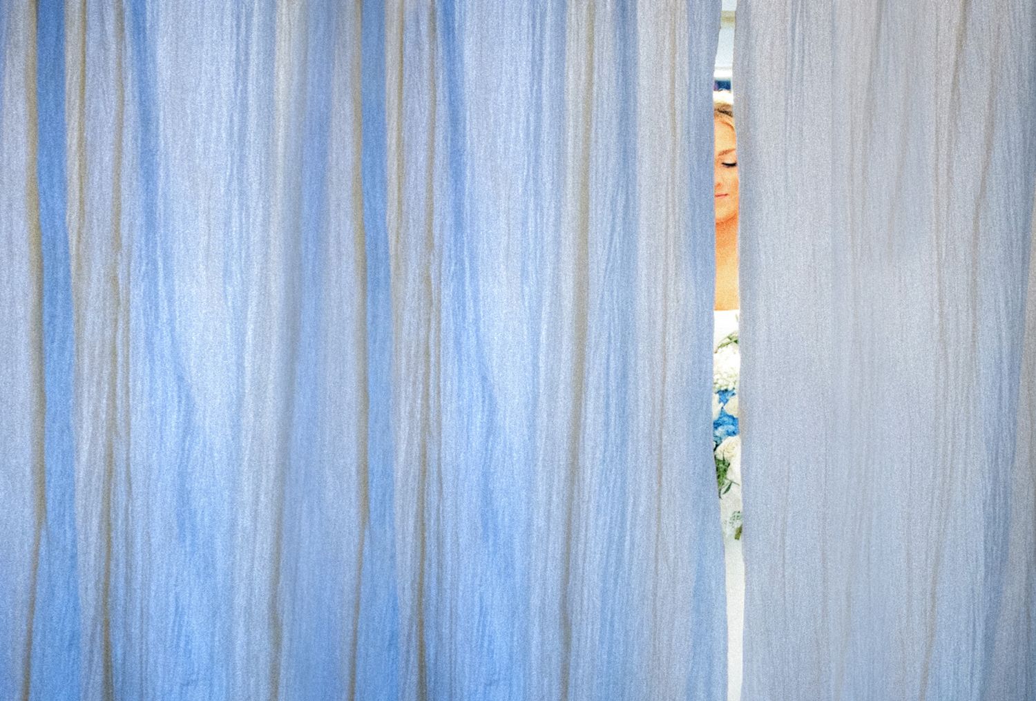 A person peeks through a gap in sheer blue curtains, partially hidden behind the flowing fabric.