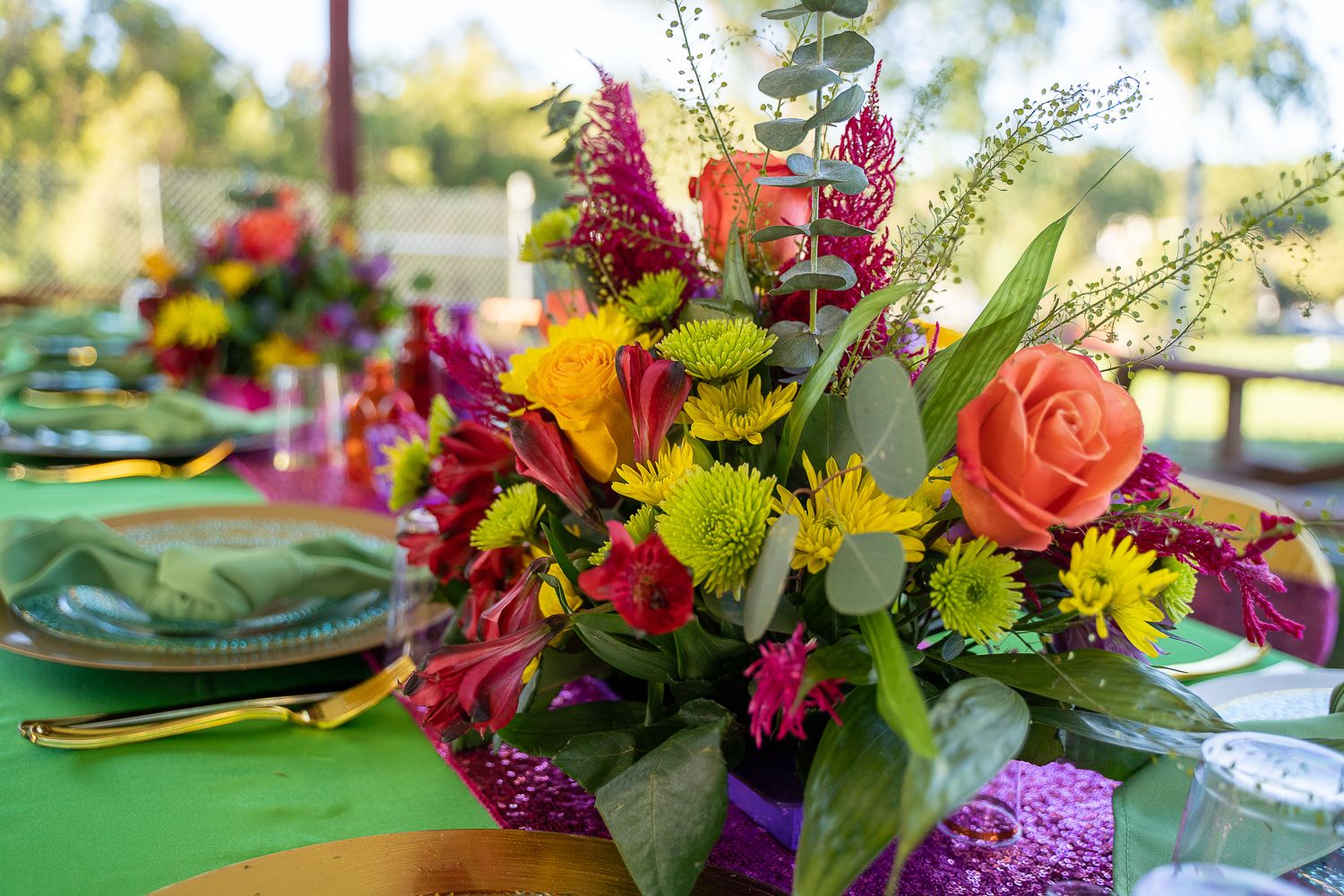 Vibrant mixed floral centerpiece with orange roses, yellow daisies, and purple blooms on a green table.