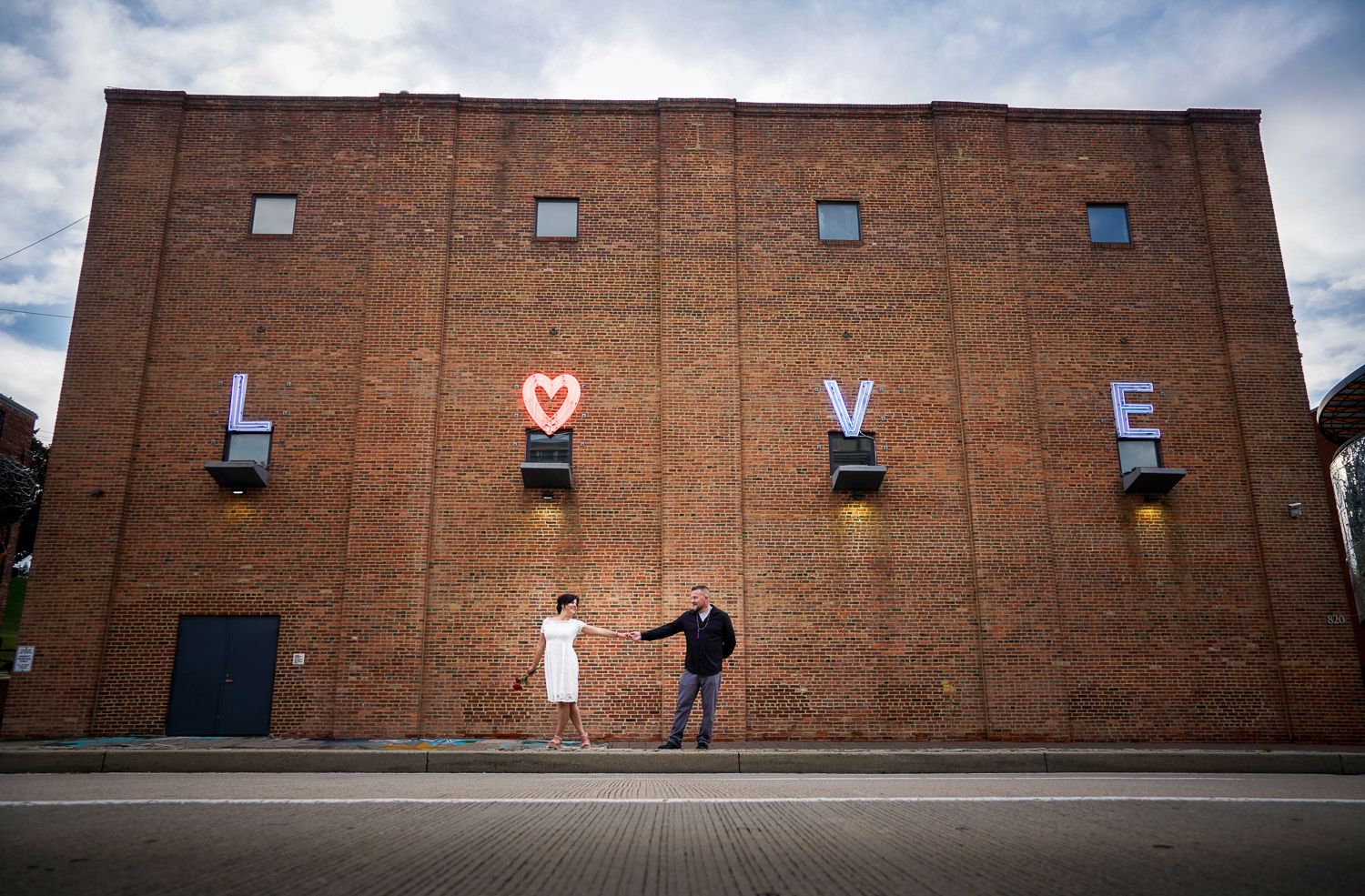 Couple holding hands in front of a large brick building decorated with illuminated LOVE letters outdoors.