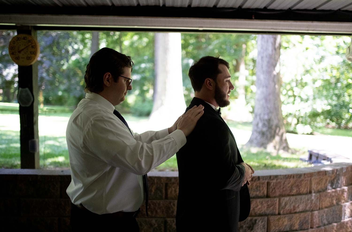 Groomsman helping groom adjust suit jacket in outdoor pavilion before wedding ceremony.