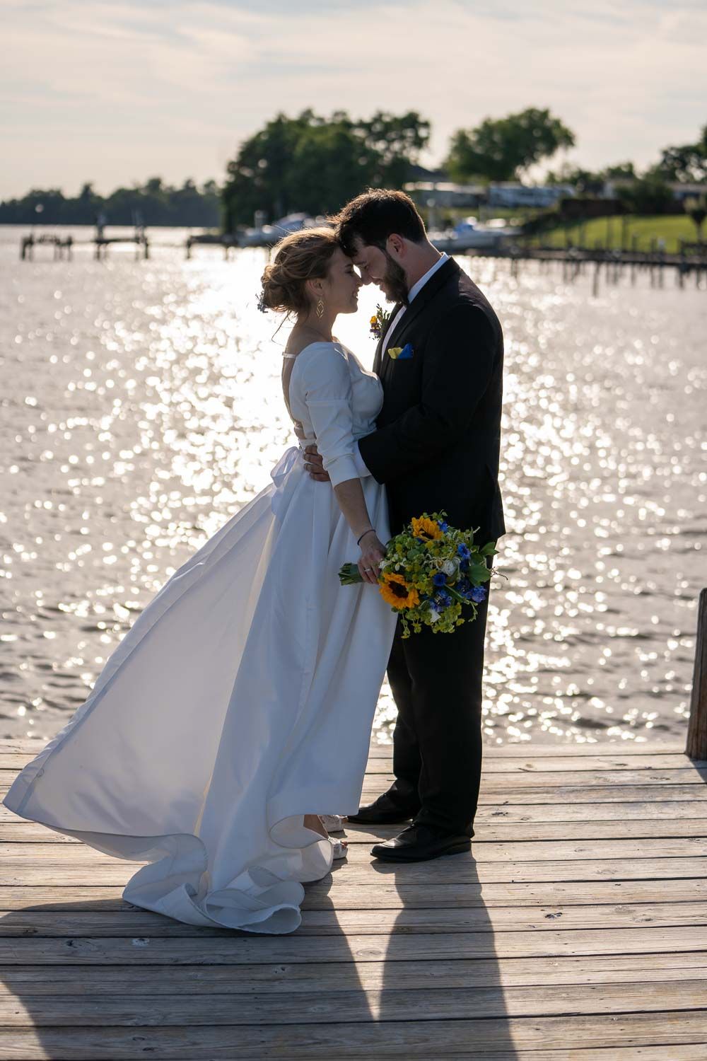 Bride and groom touching foreheads tenderly on wooden dock with shimmering water backdrop at sunset.
