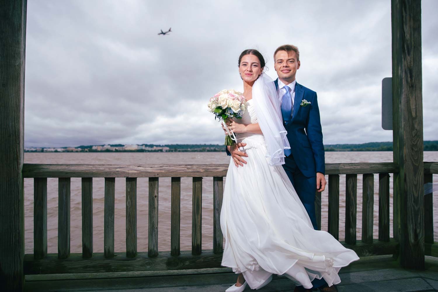 Bride and groom posing on a waterfront deck with a plane flying overhead.