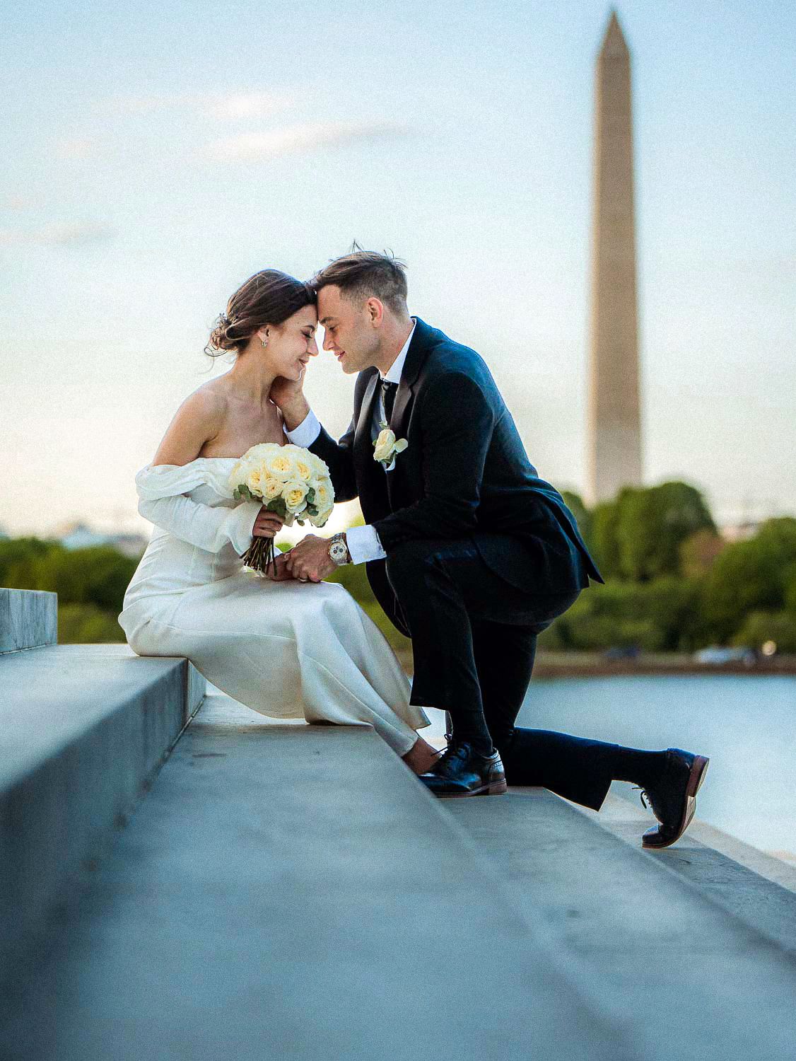 Bride and groom share an intimate moment on stone steps with the Washington Monument at dusk.