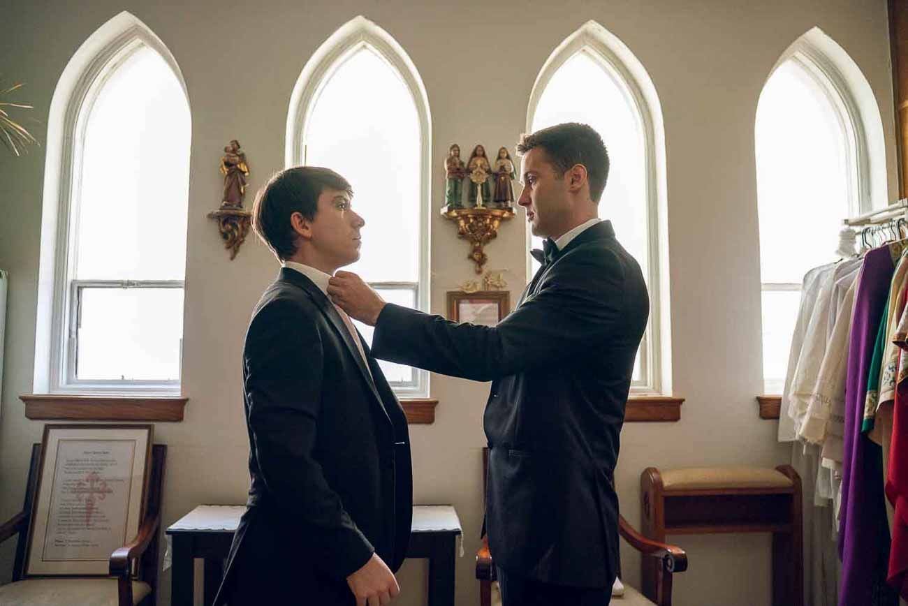 Two groomsmen in black tuxedos prepare for the wedding inside a church with arched windows.