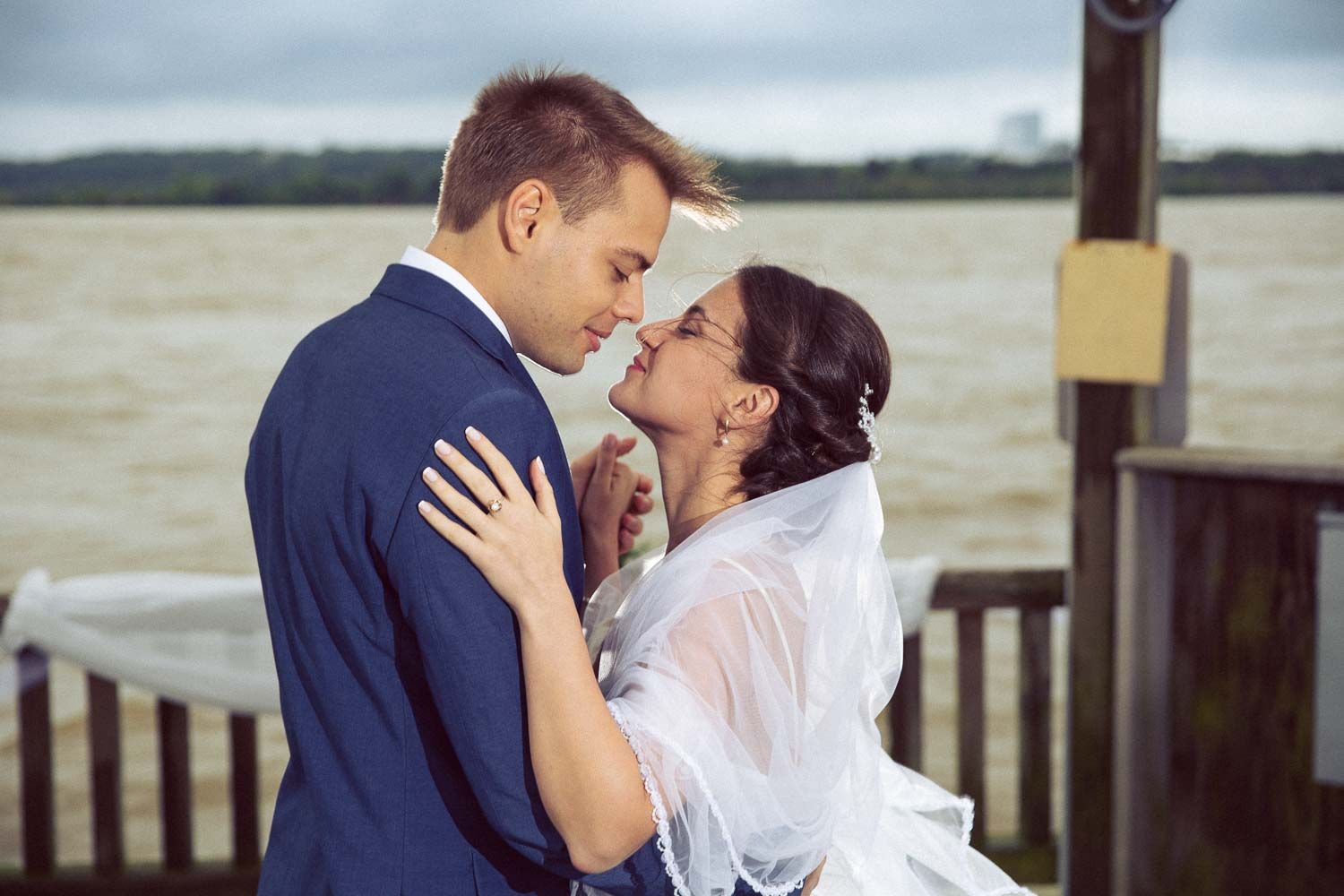 Newlyweds share tender romantic moment embracing on waterfront dock under cloudy skies.