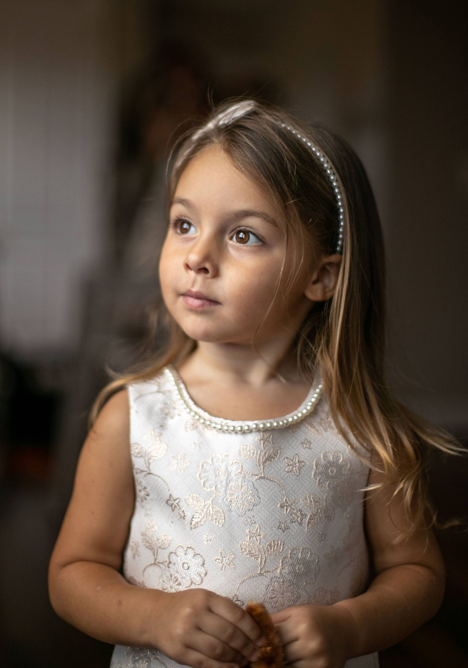 A young flower girl in a white dress with pearl headband gazes upward thoughtfully.