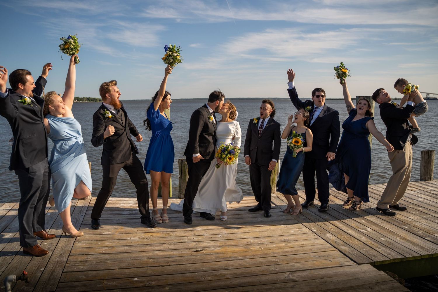Wedding party celebrates on waterfront dock raising bouquets while bride and groom kiss at sunset.