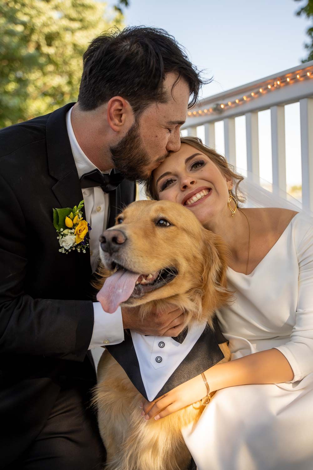Bride and groom kissing and hugging their golden retriever dressed in a tuxedo on their wedding day.