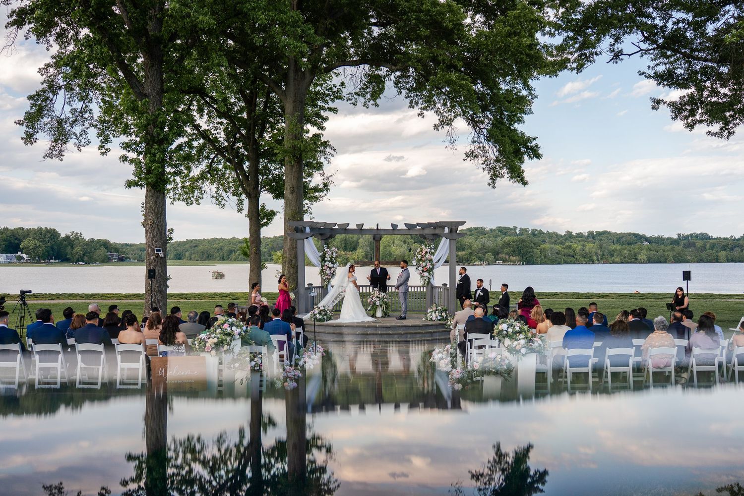 Outdoor lakeside wedding ceremony with white chairs, floral arch, and guests seated among tall trees reflected in calm water.