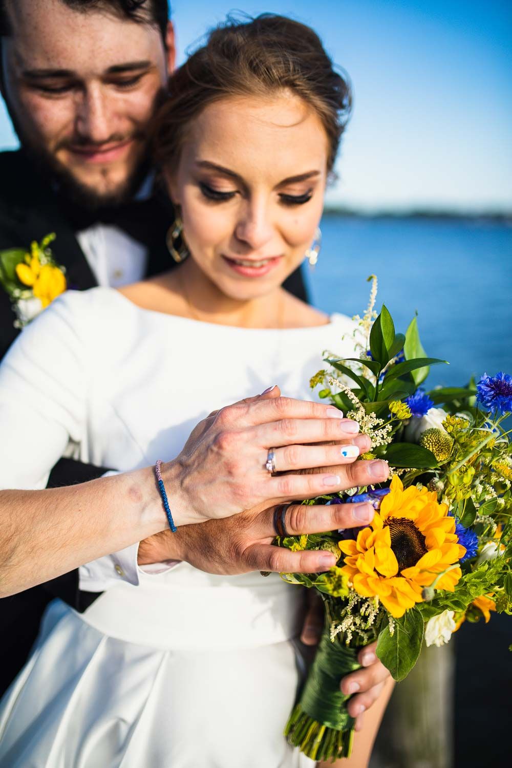 Bride and groom display wedding rings over a colorful sunflower bouquet on a waterfront dock.