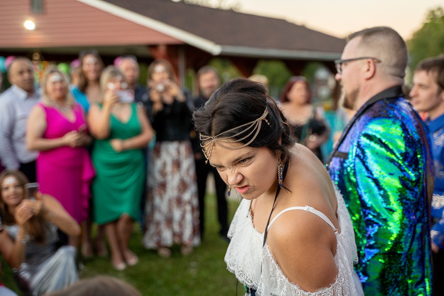 Bride in white lace dress with gold headpiece bows head before smiling wedding guests at outdoor ceremony.