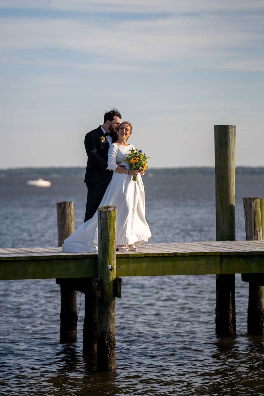 Bride and groom posing on wooden dock surrounded by water, bride carrying bright sunflower bouquet.