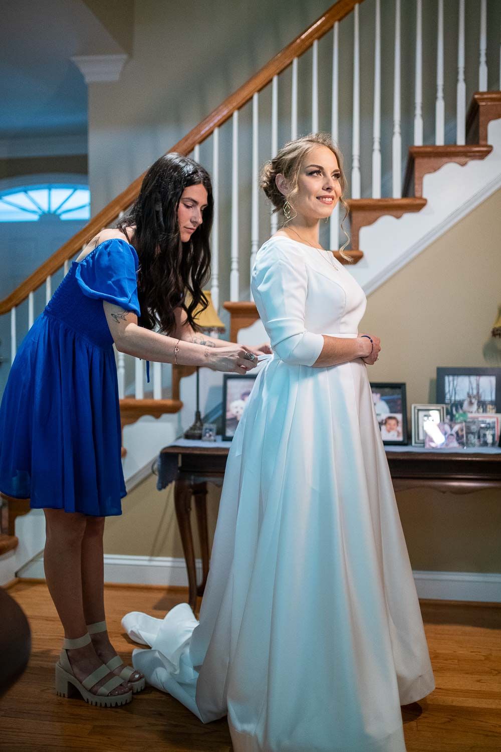 Bridesmaid in blue dress helping bride button white wedding gown near elegant staircase.