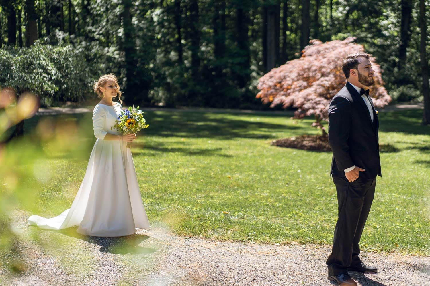 Bride in a white gown approaching groom from behind in a sunlit garden during first look moment.