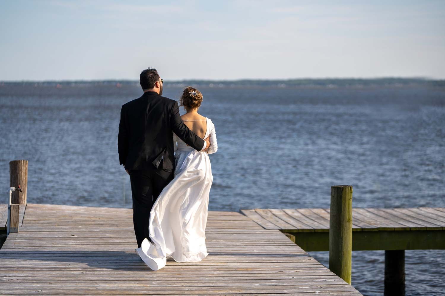 Bride and groom walking hand in hand on wooden dock overlooking calm blue water.