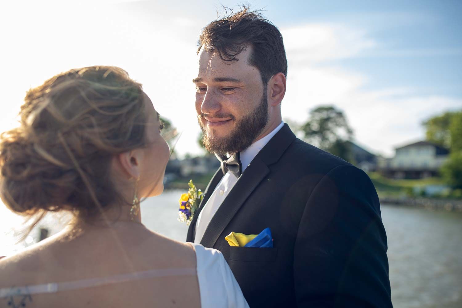 Bearded groom in tuxedo smiles warmly at bride during outdoor waterfront wedding photo session.