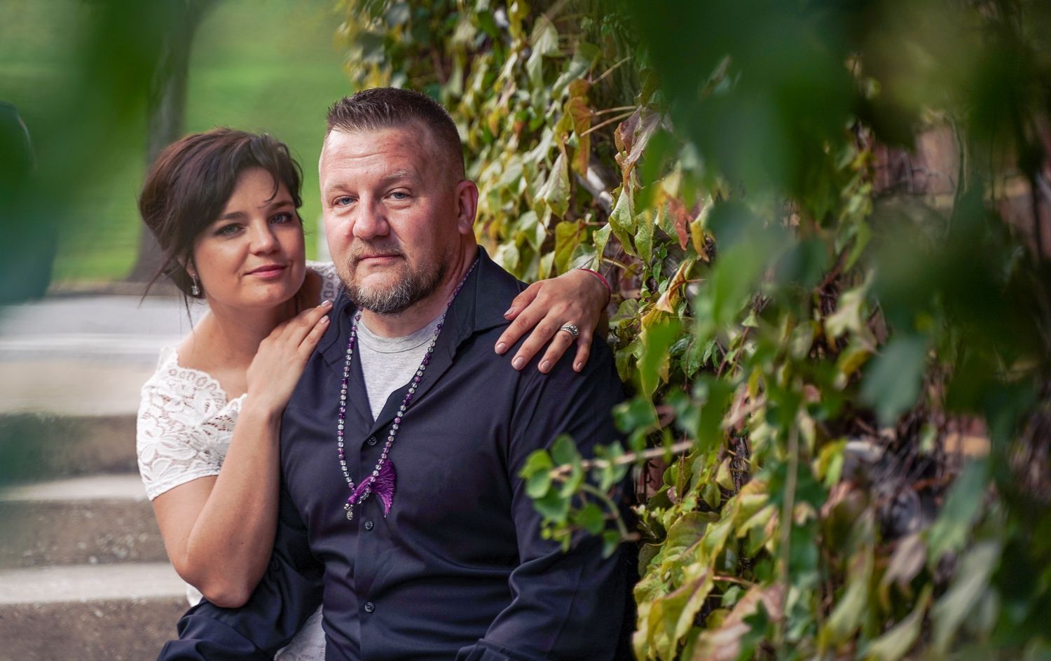 Elegant couple posing together against an ivy-covered wall, woman in white lace dress with man in vest.