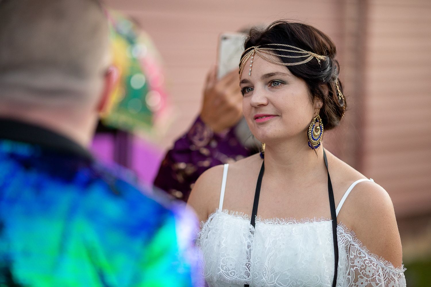Beautiful bride in white dress with headchain jewelry smiles as someone photographs her at colorful outdoor wedding.