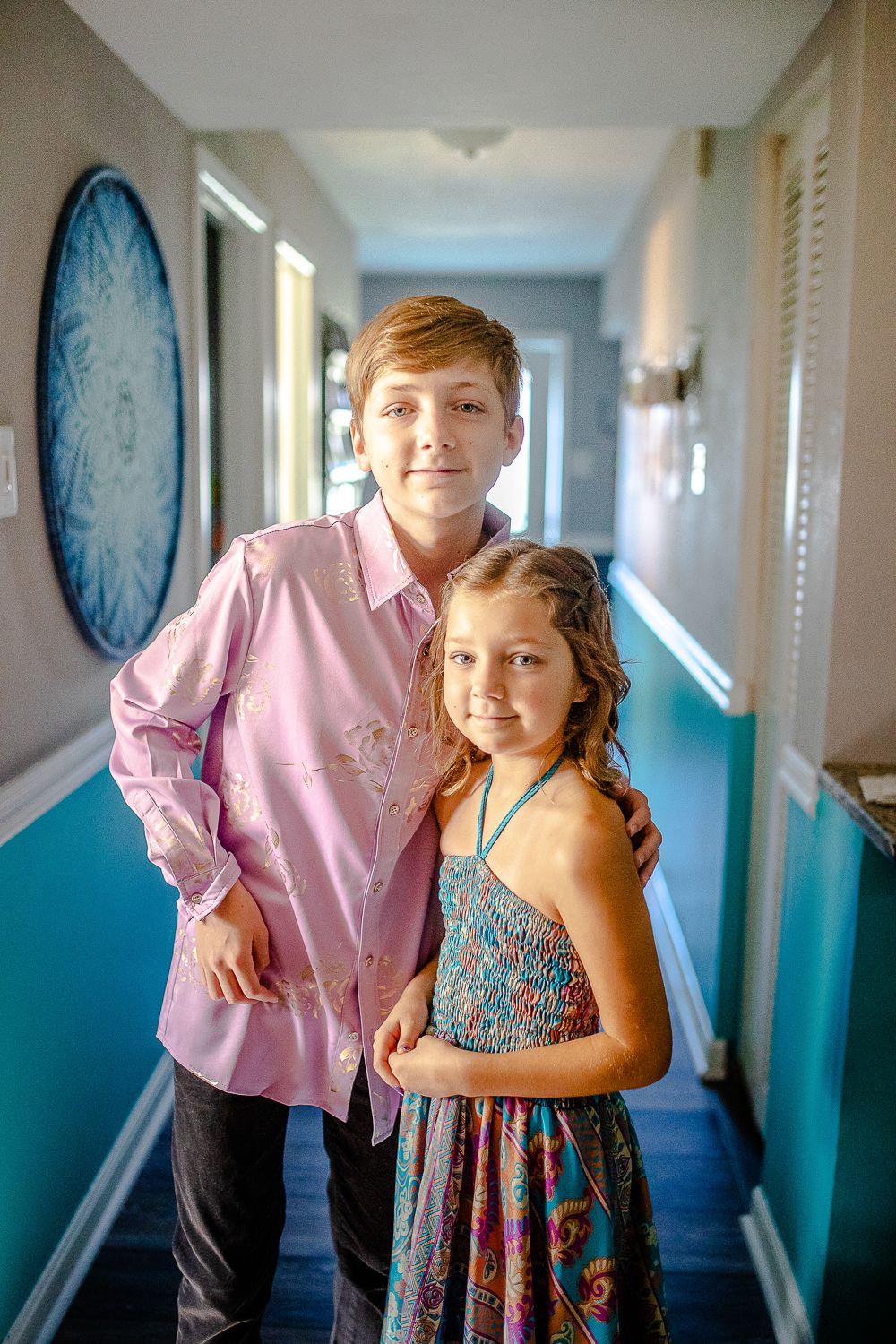 Two children, a boy in pink shirt and girl in floral dress, posing in hallway.