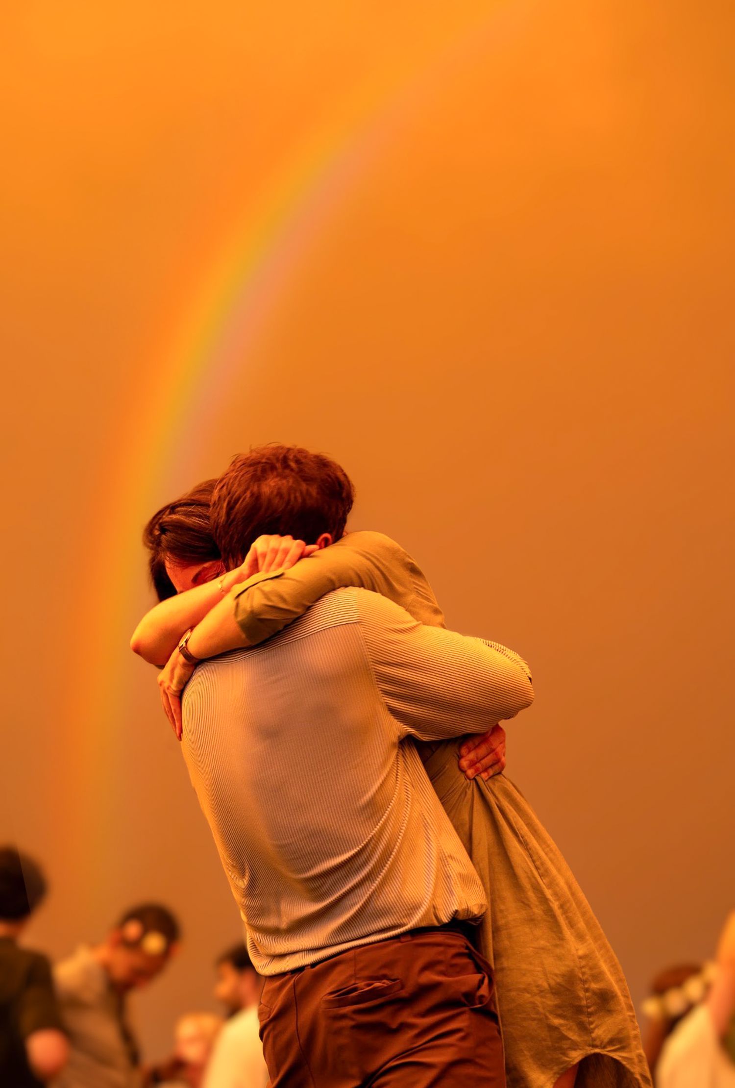 Two people embracing under a vibrant rainbow in an orange-hued sky, surrounded by a crowd.