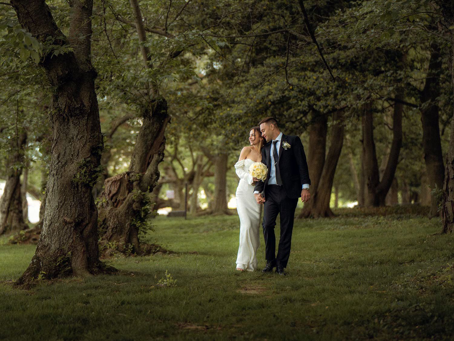 Bride and groom stroll romantically through a scenic wooded area with tall green trees.