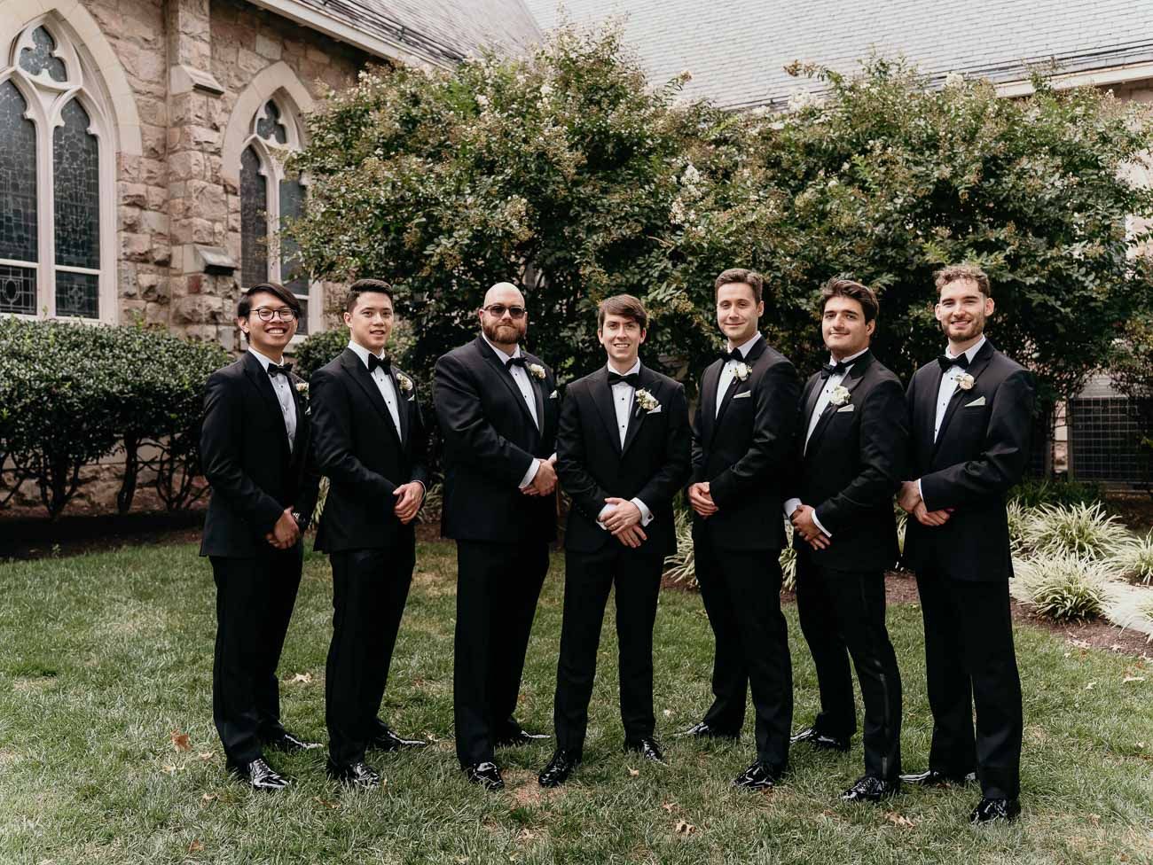 Seven groomsmen in black tuxedos with bow ties pose together outdoors near a stone church.