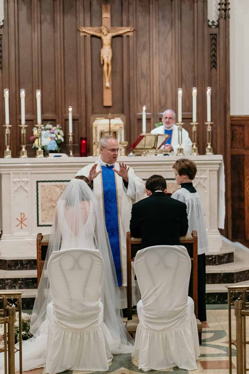 Priest officiating Catholic wedding ceremony at altar with bride in veil and groom seated in white chairs.