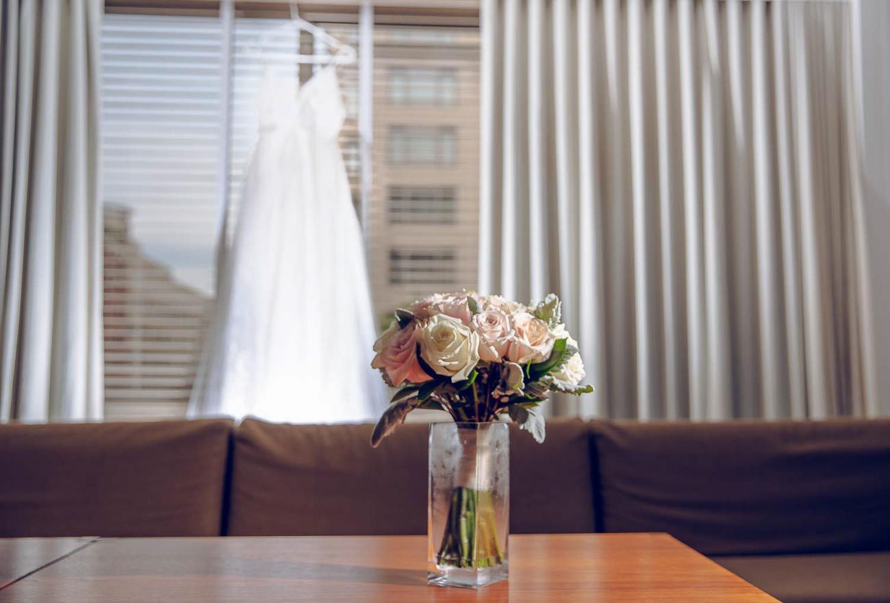 Bridal bouquet of pink and white roses in a glass vase with wedding dress hanging in a sunlit window.