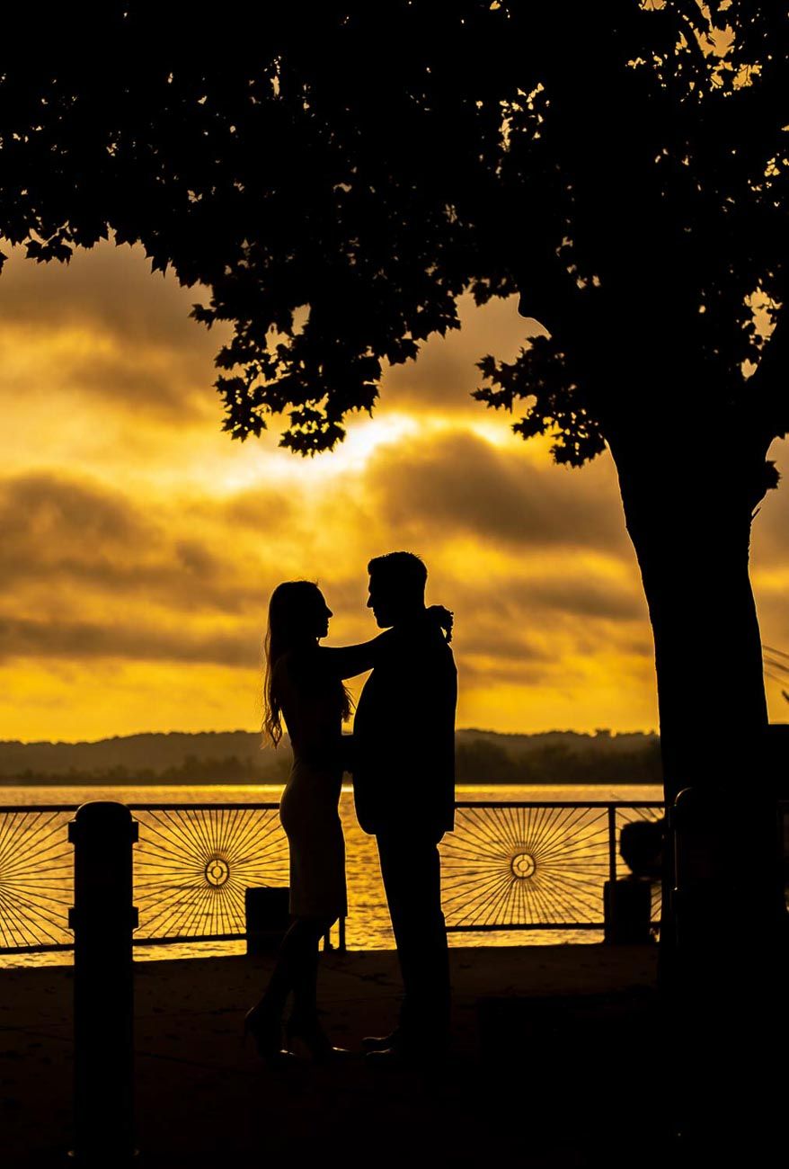 Romantic silhouette of a couple facing each other at sunset beside a shimmering waterfront.