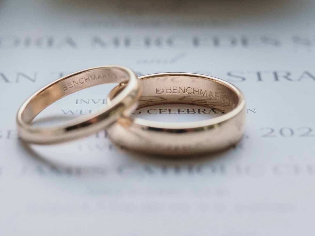 Two gold Benchmark wedding bands displayed on wedding invitation stationery.