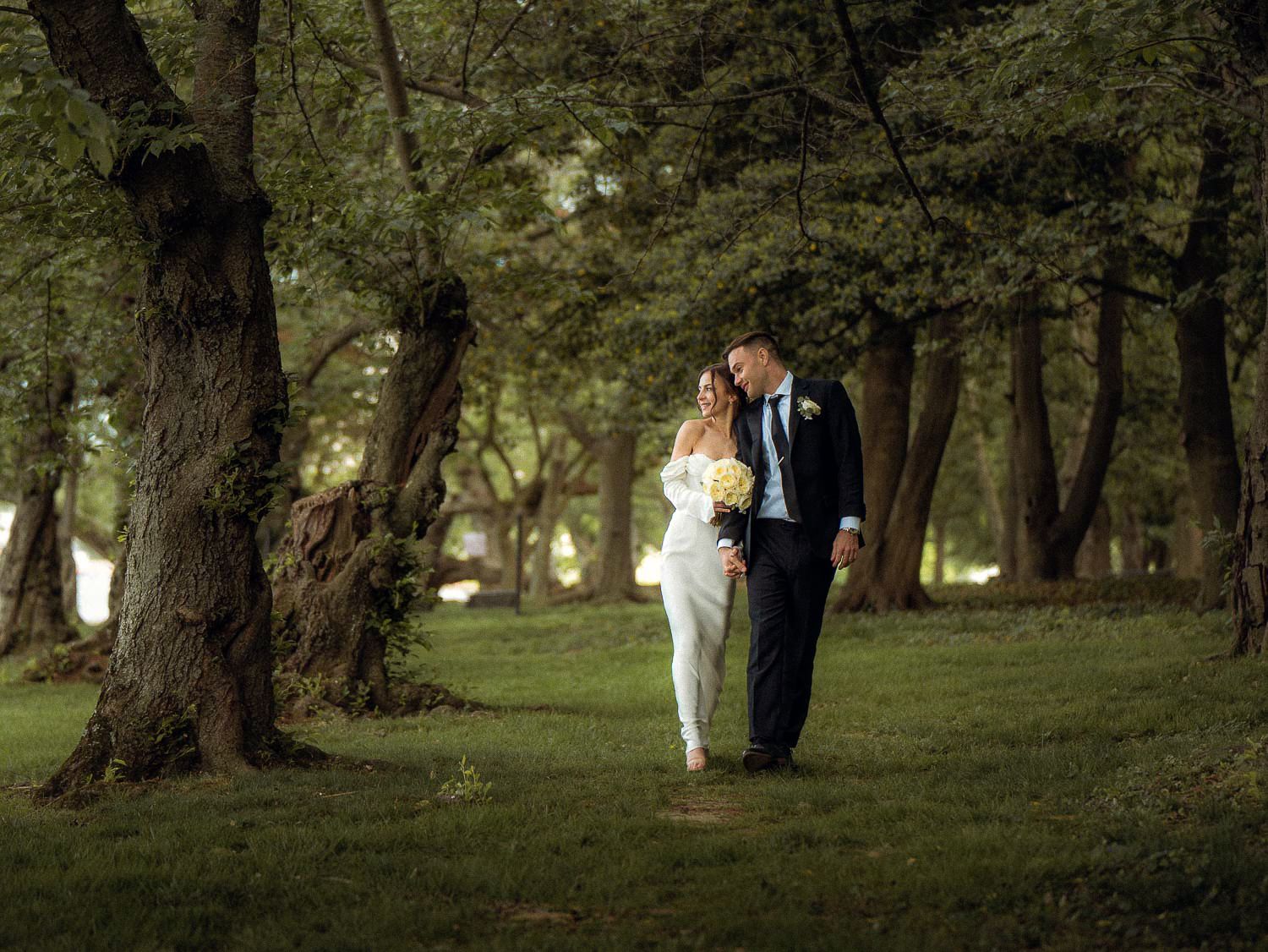 Bride and groom walk hand in hand through a sunlit forest path surrounded by tall green trees.