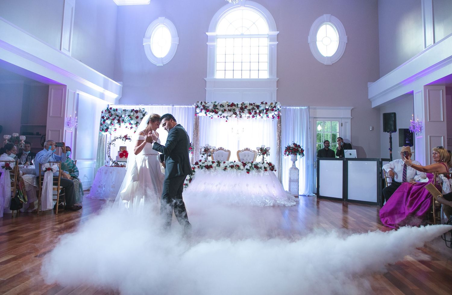A couple shares their first dance surrounded by dramatic fog on a grand ballroom dance floor.